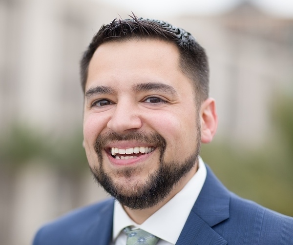 A man with short dark hair, a beard, and a navy suit smiles broadly outdoors.