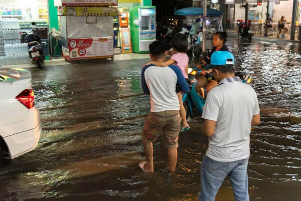 A man carries a child on his hip through a flooded urban street at night. Others walk and ride scooters in the dark water.